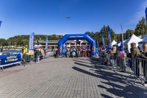Uczestnicy na brukowej alei startowej pod niebieską bramą z cyfrą 9; w niebie widać drona nad Maltą.