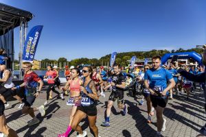 Start na bruku przy scenie: uczestnicy w biegu, w tym kobieta w różowym biustonoszu sportowym; ktoś nagrywa telefonem.