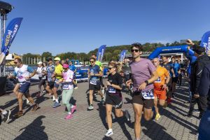 Pierwsze metry trasy: biegacze w fioletowych i różowych koszulkach na bruku z widokiem jeziora w tle.