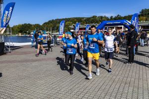 Biegacze z numerami 30, 21 i 117 mkną ku kamerze; za nimi niebieska brama nad taflą wody.