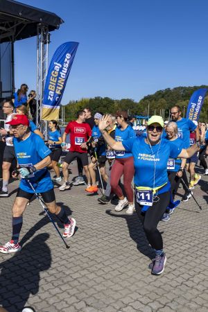Uśmiechnięta biegaczka w limonkowej czapce rozpościera ręce; w tle nordic walking i niebieskie flagi.