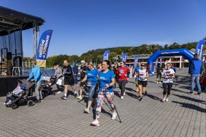 Dwie uczestniczki z kijkami nordic walking idą obok mężczyzny popychającego wózek na brukowanej alei.