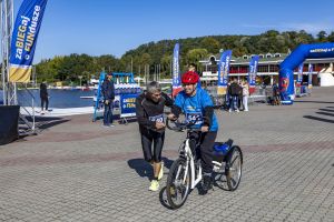 Zawodnik na trójkołowym rowerze z numerem 541; obok biegnie towarzysz w neonowych butach przy Jeziorze Maltańskim.