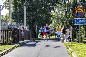 Trójka mężczyzn biegnie po asfalcie mostu; po prawej znak rzeki Cybiny i zakaz skoków do wody.