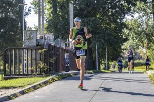 Numer 682 w zielono-czarnej koszulce biegnie wzdłuż alei wśród liściastych drzew w pełnym słońcu.