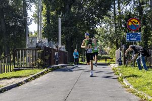 Biegacze w zielonych koszulkach z numerami 559 i 195; operator na trawie filmuje kamerą na gimbalu.