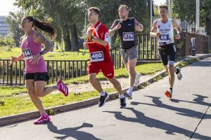 Czworo biegaczy z numerami 164, 460, 679 i 65 mknie aleją nad wodą; na trawniku gołębie.