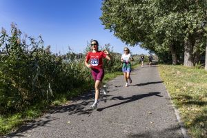 Uśmiechnięta kobieta w czerwonej koszulce z numerem 476 macha do kamery podczas biegu nad Jeziorem Maltańskim.