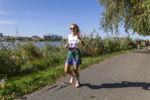 Blondynka w koszulce Run Team Rydzyna i kolorowej spódnicy biegowej pokazuje cztery palce nad brzegiem jeziora.