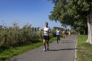 Mężczyzna w okularach i białej koszulce biegowej na czele kilku uczestników na alei nad jeziorem w pogodny dzień.
