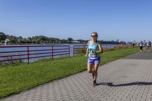 Kobieta w jasnoniebieskim topie Nike, fioletowych spodenkach i różowych butach z numerem 399 na brukowanej ścieżce.