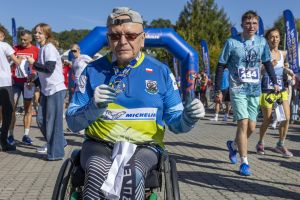 Starszy mężczyzna na wózku z logo Michelin, medalem na szyi i białą torbą, gestem kciuka w strefie finiszu.