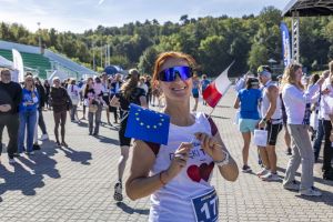 Uczestniczka z numerem 177 trzyma flagi Unii Europejskiej i Polski, uśmiechając się w okularach na brukowanym placu.