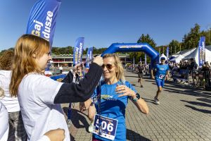 Wolontariuszka wręcza medal uśmiechniętej biegaczce z numerem 506 w niebieskiej koszulce pod bramą finiszu.