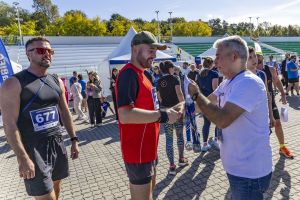 Uczestnik w czerwonej koszulce i zielonej czapce odbiera medal i ściska dłoń organizatora; obok biegacz z numerem 677.