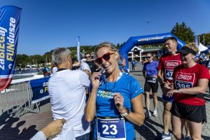 Biegaczka z numerem 23 w niebieskiej koszulce trzyma medal finishera przy ustach na tle Jeziora Malty.