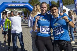 Biegaczka z numerem 380 i biegacz z numerem 387 uśmiechają się do obiektywu, unosząc medale pod niebieską bramą.