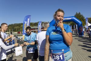 Blondynka z numerem 381 gryzie medal finishera w niebieskiej koszulce na tle bramy i wolontariuszy.