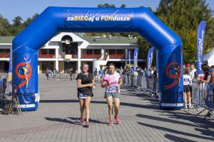 Dwie biegaczki z numerami 599 i 672 biegną razem pod niebieską bramą przy białym budynku regatowym.