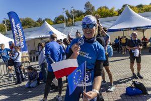 Blondynka z numerem 8 unosi medal oraz flagi Polski i Unii Europejskiej przy białych namiotach eventu.