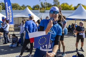 Uczestniczka z numerem 118 przygryza medal, trzymając flagi Polski i UE przy namiotach na Malcie.
