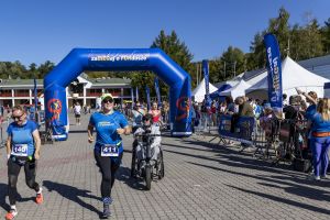 Uczestniczki z numerami 411 i 140 oraz zawodnik na wózku sportowym biegną pod niebieską bramą startu.