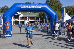 Kobieta z numerem 116 w żółtej saszetce biegnie pod niebieską bramą z napisem zaBIEGaj o FUNdusze.