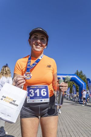 Uśmiechnięta biegaczka z numerem 316 trzyma papierową torbę i butelkę wody z medalem na szyi.