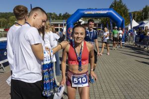 Biegaczka w koralowej bluzce z numerem 510 z medalem stoi obok wolontariusza z wstążkami nagród.