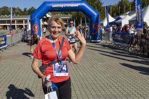Biegaczka w czerwonej koszulce z numerem 74 macha ręką, trzymając torbę i butelkę przy bramie mety.