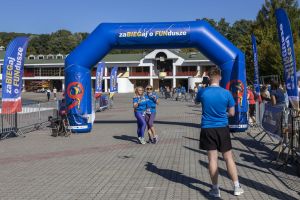 Dwie biegaczki z numerami 561 i 828 pozują pod bramą, mężczyzna w pierwszym planie robi im zdjęcie telefonem.