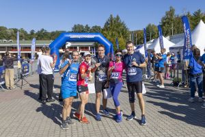Pięcioro uczestników z medalami pozuje przed niebieską bramą mety w słoneczny dzień biegu.