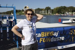 Organizatorka w koszulce zaBIEGaj o FUNdusze stoi przy stojaku z medalami i banerem nad jeziorem Maltańskim.
