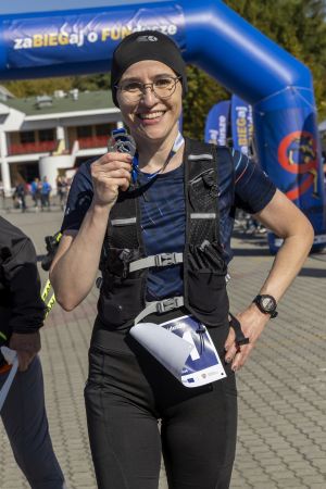 Biegaczka w kamizelce hydracyjnej unosi srebrny medal finishera przed niebieską bramą mety.