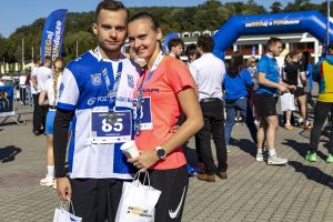 Para finisherów z medalami przy niebieskiej bramie mety; mężczyzna z numerem 85 i białą torbą prezentową.