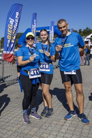 Trójka uczestników w niebieskich koszulkach z medalami; numery 411 i 418, w tle banery i brama mety.