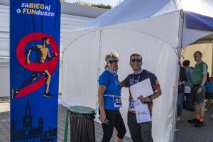 Para z medalami przy wysokim banerze biegu; kobieta z numerem 55, mężczyzna z torbą i białym pakietem.