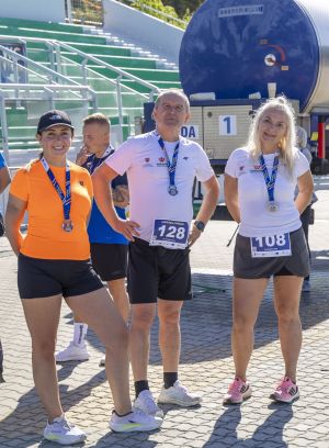 Trójka finisherów z medalami na bruku; numery 128 i 108, kobieta w pomarańczowej koszulce i czapce The North Face.