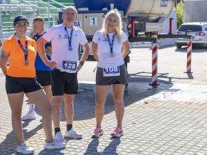 Finisherzy z numerami 128 i 108 przy niebieskim zbiorniku BOROWSKI; trybuny i samochód w tle na słońcu.
