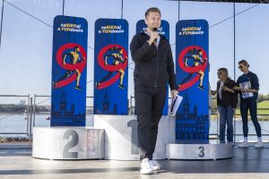 Prowadzący z mikrofonem przy srebrnym podium 1–2–3; wysokie banery biegu i Jezioro Maltańskie za sceną.