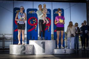 Dekoracja na podium: trzy biegaczki z żółtymi kopertami nagród, zwyciężczyni na środku trzyma dziecko.