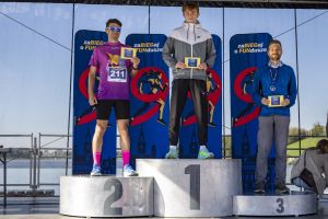 Trzech mężczyzn na podium z dyplomami nad Maltą; środkowy w kurtce Nike, po lewej numer 211, po prawej medal.