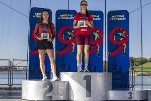 Dwie laureatki na podium z żółtymi kartami nagrody; trzeci stopień pusty, banery biegu i tafla Malty w tle.