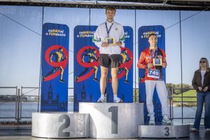 Dwóch mężczyzn na podium — pierwsze i trzecie miejsce; zwycięzca w białej kurtce Adidas, numer 749 na stopniu trzecim.