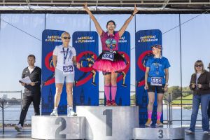 Trzy laureatki na podium: zwyciężczyni w różowym tutu unosi ręce w geście radości, obok druga i trzecia z medalami.