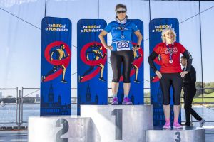 Dwie biegaczki z medalami stoją na stopniach 1 i 3 podium; w tle tafla Jeziora Maltańskiego i banery imprezy.