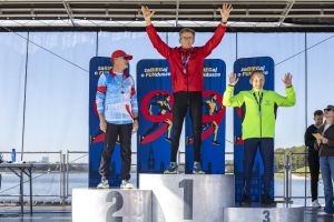 Trzech mężczyzn na podium: zwycięzca w czerwonej kurtce wznosi ręce, obok drugie i trzecie miejsce z medalami.