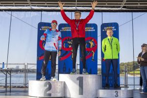 Podium z trzema biegaczami nad jeziorem; środkowy laureat świętuje z uniesionymi ramionami w słońcu.