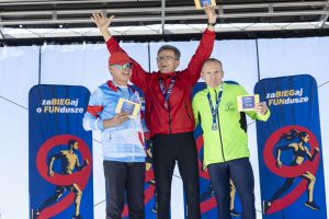 Ceremonia na scenie: trzech biegaczy z medalami i dyplomami, zwycięzca w czerwonej bluzie w geście triumfu.