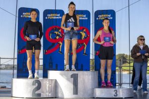 Trzy biegaczki na srebrnym podium z niebieskimi kopertami nagród; za nimi woda Malti i banery zawodów.